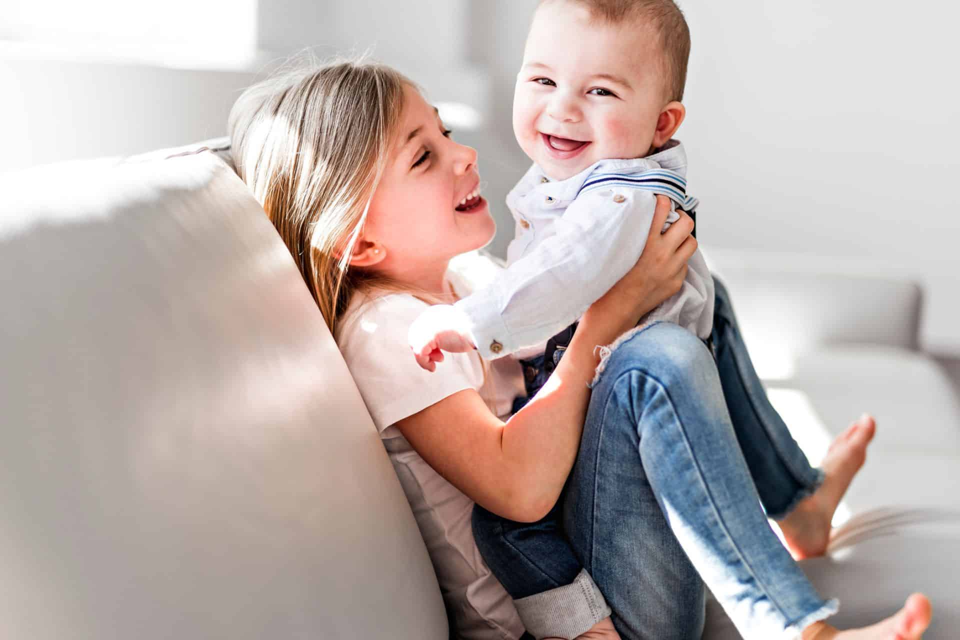 Young girl holding a smiling baby while sitting on a couch.